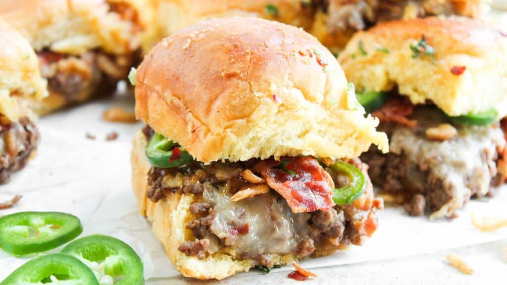 Close-up of a slider sandwich with ground beef, melted cheese, bacon, and jalapeño slices on a soft bun, surrounded by more sliders and jalapeño slices.