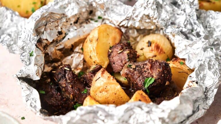 Cooked beef chunks and potato pieces with herbs in an open foil packet.