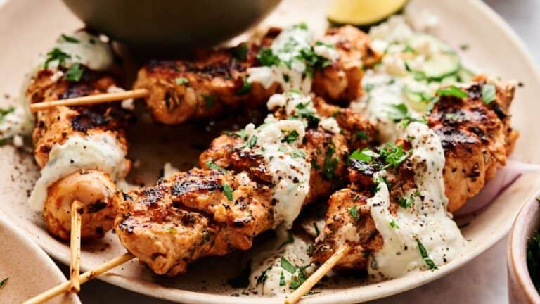 Grilled chicken skewers topped with a white sauce and chopped herbs are served on a plate with sliced vegetables and a lemon wedge.
