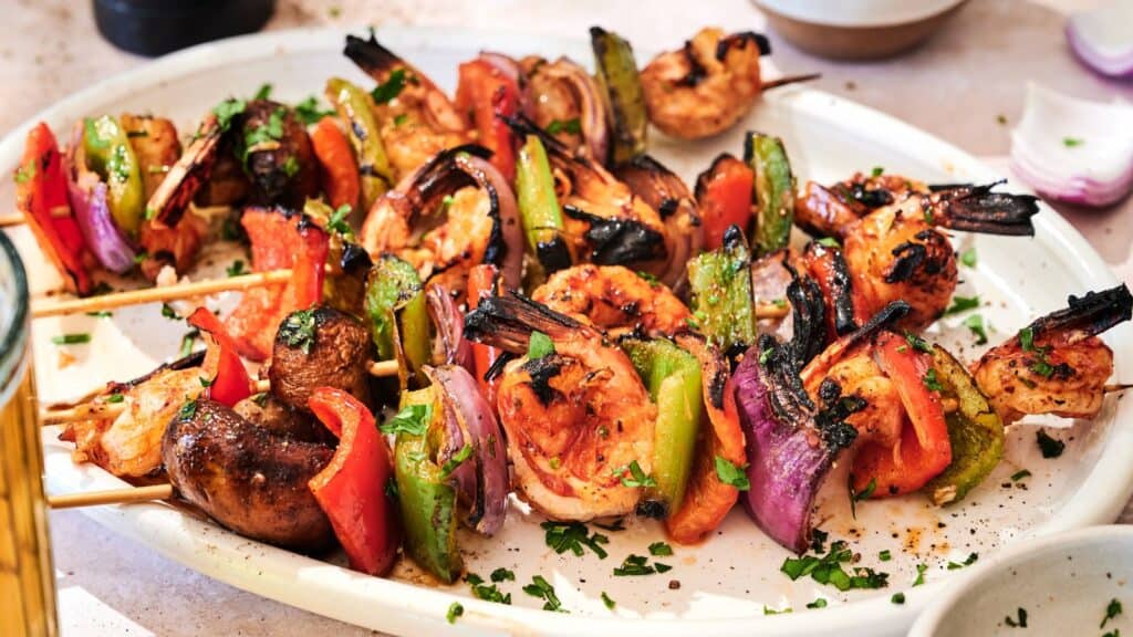 Grilled shrimp and vegetable skewers with mushrooms, bell peppers, red onion, and herbs on a white platter.