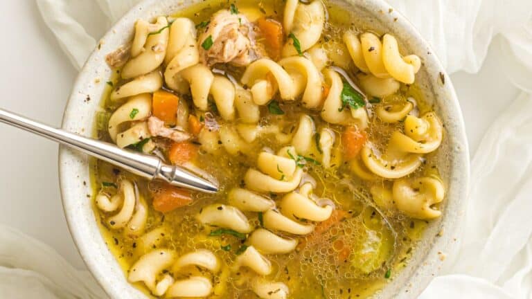 A bowl of noodle soup with rotini pasta, chunks of chicken, carrots, celery, and herbs, shown with a spoon resting inside.