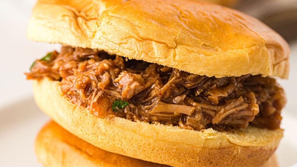A close-up of a pulled pork sandwich with shredded meat in barbecue sauce served on a soft bun.
