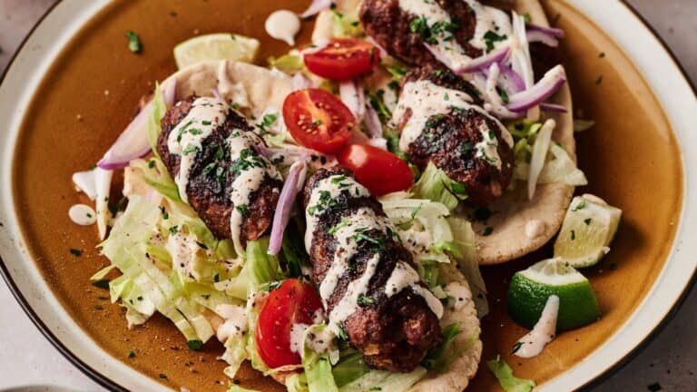 Grilled meat skewers on pita bread with shredded lettuce, cherry tomatoes, sliced red onions, lime wedges, and a drizzle of white sauce on a brown plate.