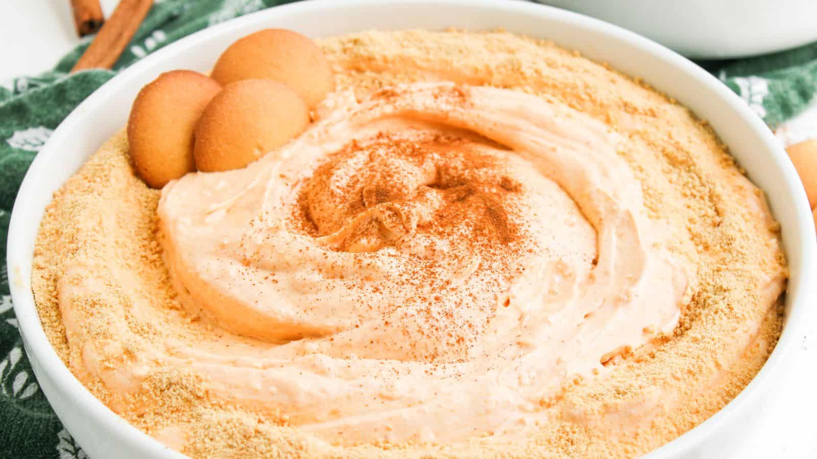 A bowl of creamy dip topped with crushed cookies and a dusting of cinnamon, garnished with whole vanilla wafers on the side.