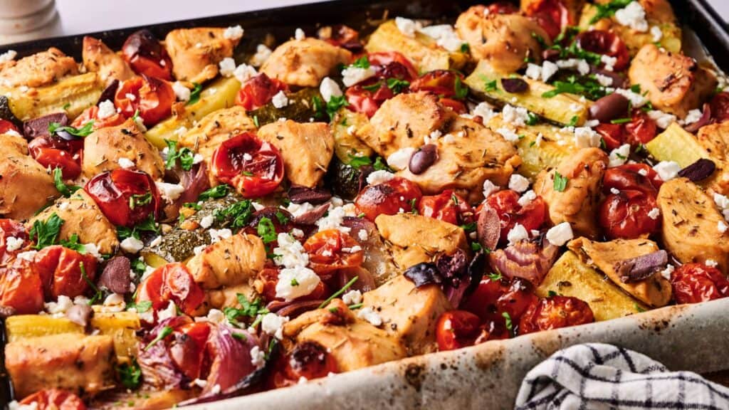 A baking sheet filled with baked chicken, cherry tomatoes, zucchini, olives, feta cheese, and herbs, arranged in a colorful, roasted medley.