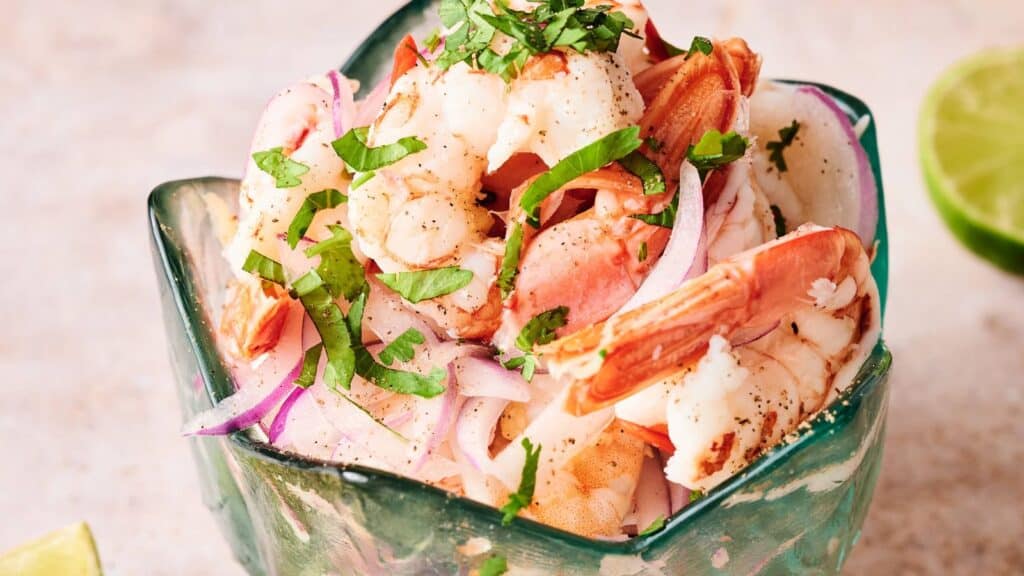 A glass bowl filled with shrimp, sliced red onions, and chopped cilantro, with a lime half visible in the background.