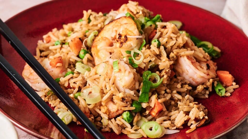 A red plate of shrimp fried rice garnished with sliced green onions and carrots, served with black chopsticks.