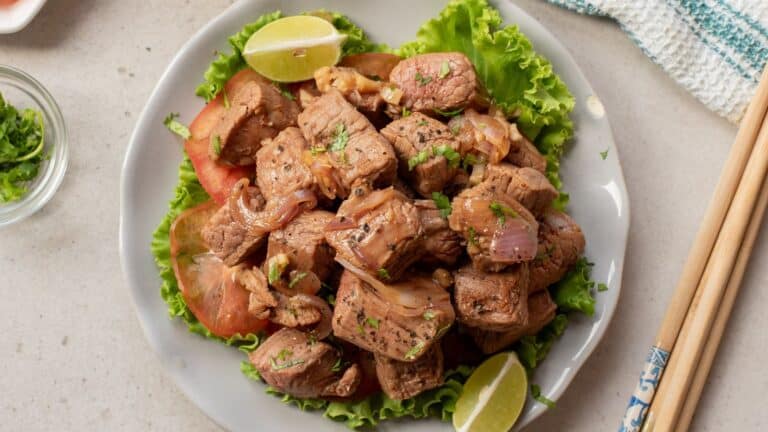 A plate of sautéed beef cubes with onions, served on lettuce and tomato slices, garnished with lime wedges and herbs. Chopsticks rest beside the plate.