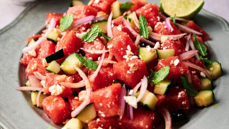 A plate of watermelon salad with cucumber, red onion, feta cheese, and fresh mint leaves, garnished with a lime wedge.