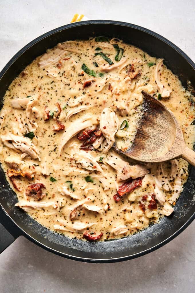 A skillet filled with creamy Marry Me Chicken Pasta Salad, sun-dried tomatoes, and herbs is being stirred with a wooden spoon.