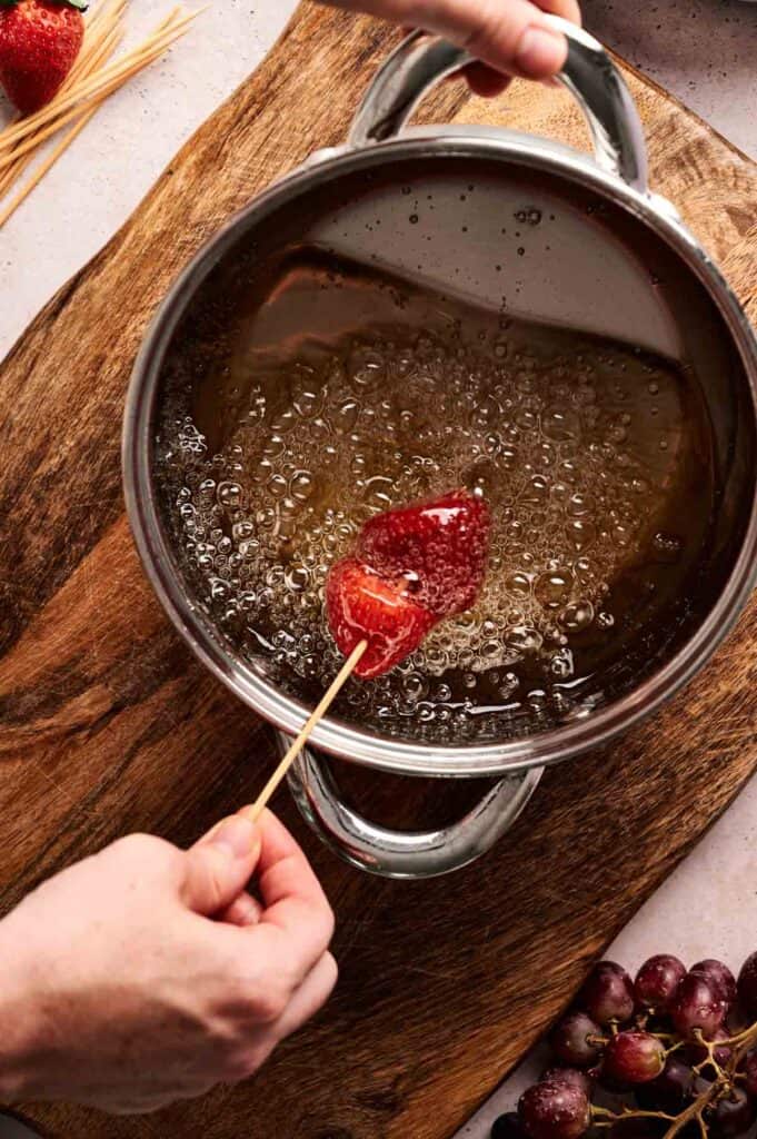 A hand dips a strawberry on a skewer into bubbling sugar syrup in a saucepan, with grapes and uncooked skewers nearby on a wooden surface.