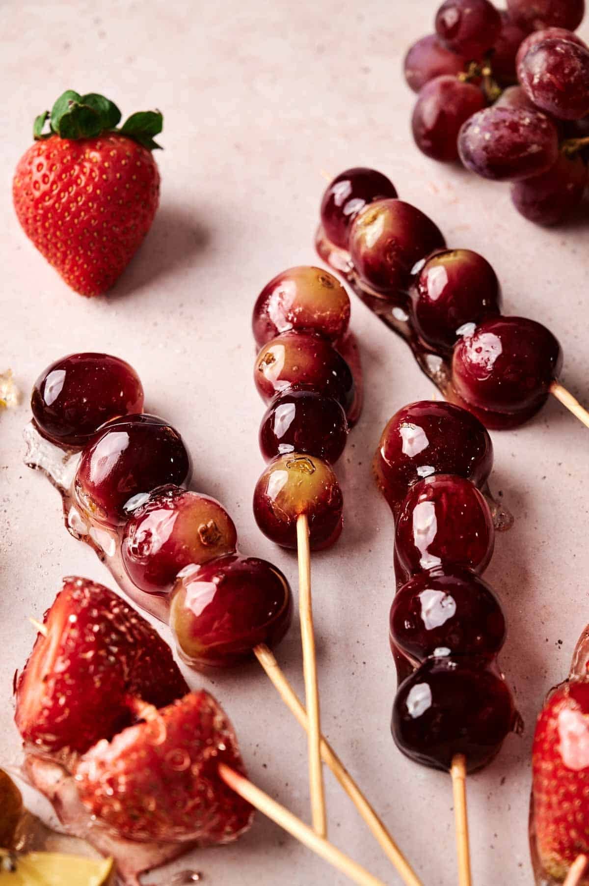 Three skewers with candied red grapes and two with candied strawberries are on a light surface, surrounded by loose grapes and a whole strawberry.