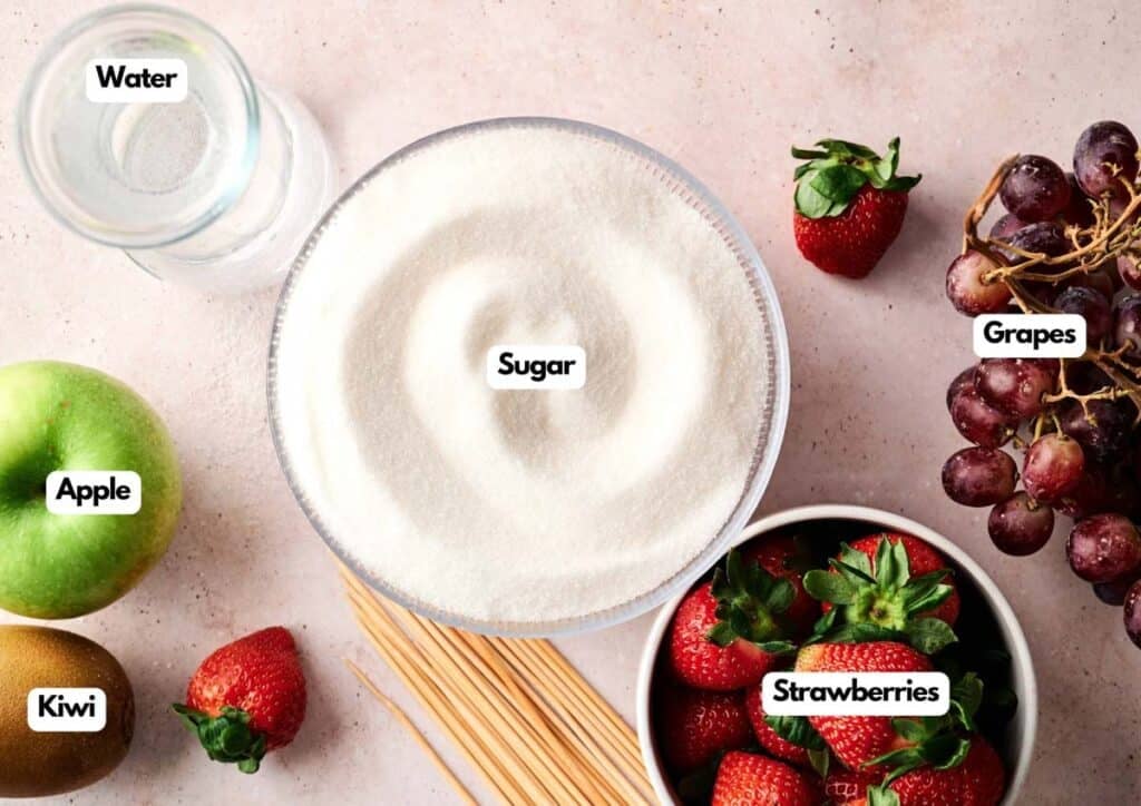 Flat lay of Tanghulu ingredients for a recipe, featuring a bowl of sugar, water, apple, kiwi, grapes, strawberries, and uncooked spaghetti, each clearly labeled.