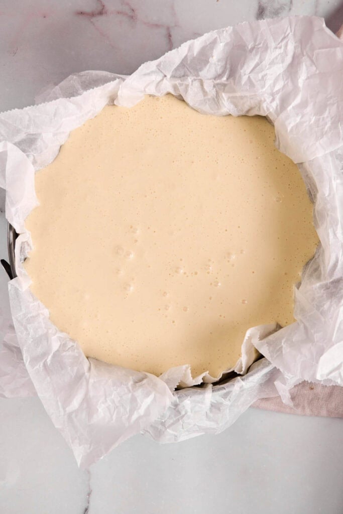 A round baking pan lined with parchment paper is filled with smooth, pale basque cheesecake batter, ready to be baked.