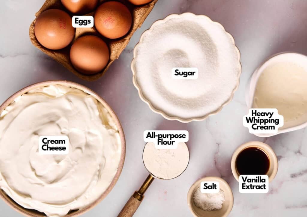 Top-down view of labeled baking ingredients for basque cheesecake: eggs, sugar, heavy whipping cream, cream cheese, all-purpose flour, salt, and vanilla extract on a marble surface.