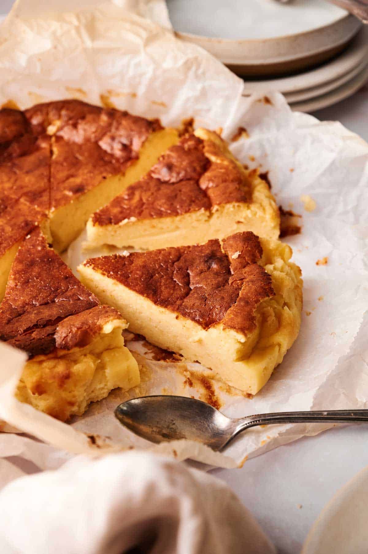 A sliced Basque cheesecake rests on parchment paper with a serving spoon beside it and stacked plates in the background.