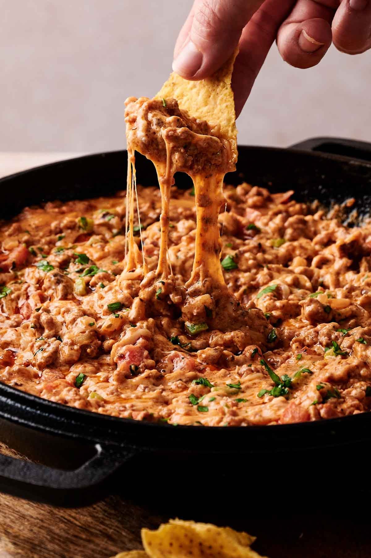 A hand dips a tortilla chip into a skillet of cowboy queso dip, lifting out a portion of melted cheese and ground meat with gooey cheese stretching.