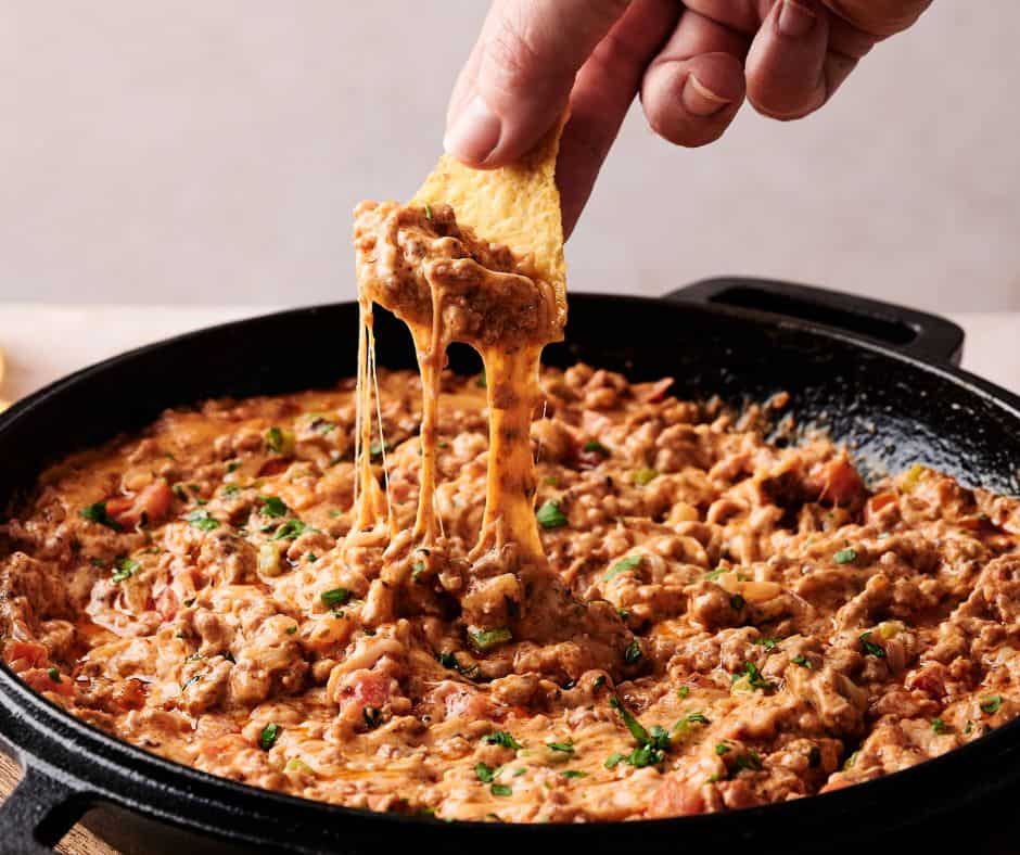 A hand dips a tortilla chip into a skillet of cowboy queso dip, bursting with cheesy, meaty goodness, herbs, and tomato pieces.