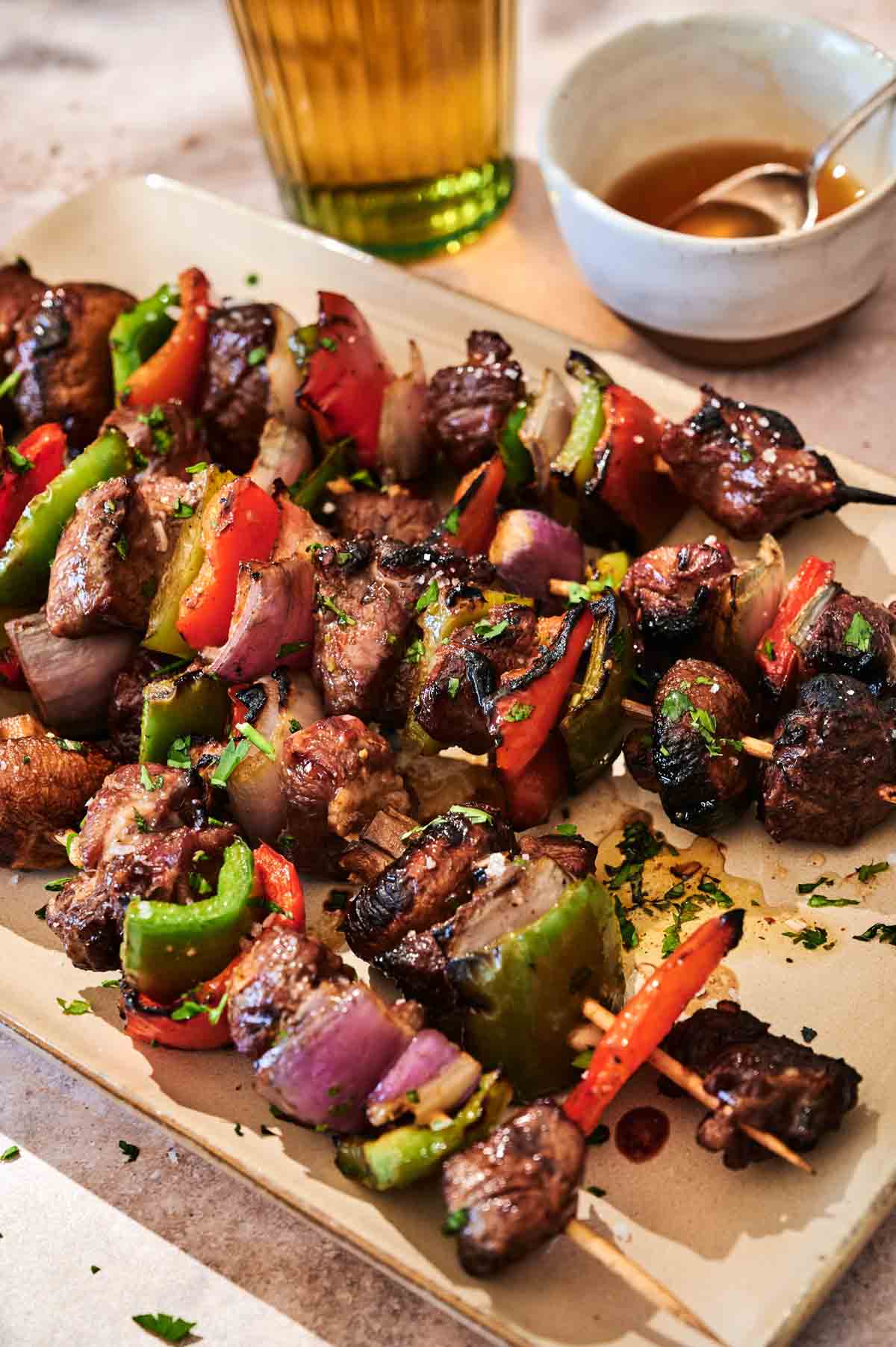 Grilled Steak Kabobs with red onion, bell peppers, and mushrooms are served on a rectangular platter, accompanied by a sauce bowl and a refreshing drink in the background.
