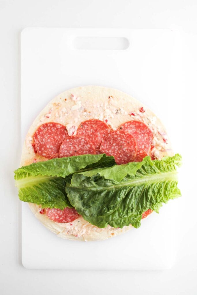 A tortilla wrap on a white cutting board topped with a creamy spread, slices of salami, and romaine lettuce leaves&mdash;perfect for making delicious Italian Sub Pinwheels.