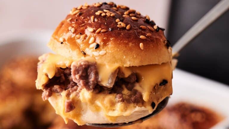 A close-up of a cheeseburger slider with ground beef and melted cheese on a sesame seed bun, being lifted with a metal spatula.