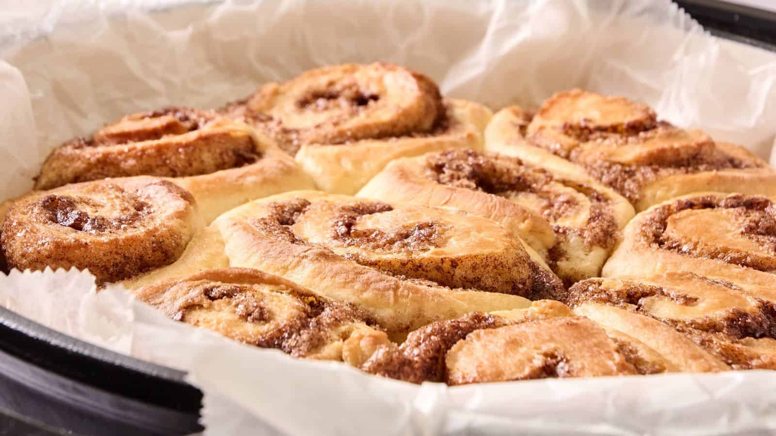A close-up of freshly baked cinnamon rolls in a parchment-lined baking dish, showing golden-brown swirls and a soft texture.