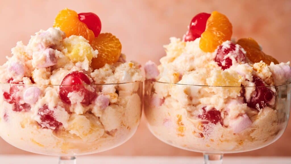 Two glass bowls filled with ambrosia salad, containing whipped mixture, cherries, mandarin oranges, pineapple, and marshmallows against a neutral background.
