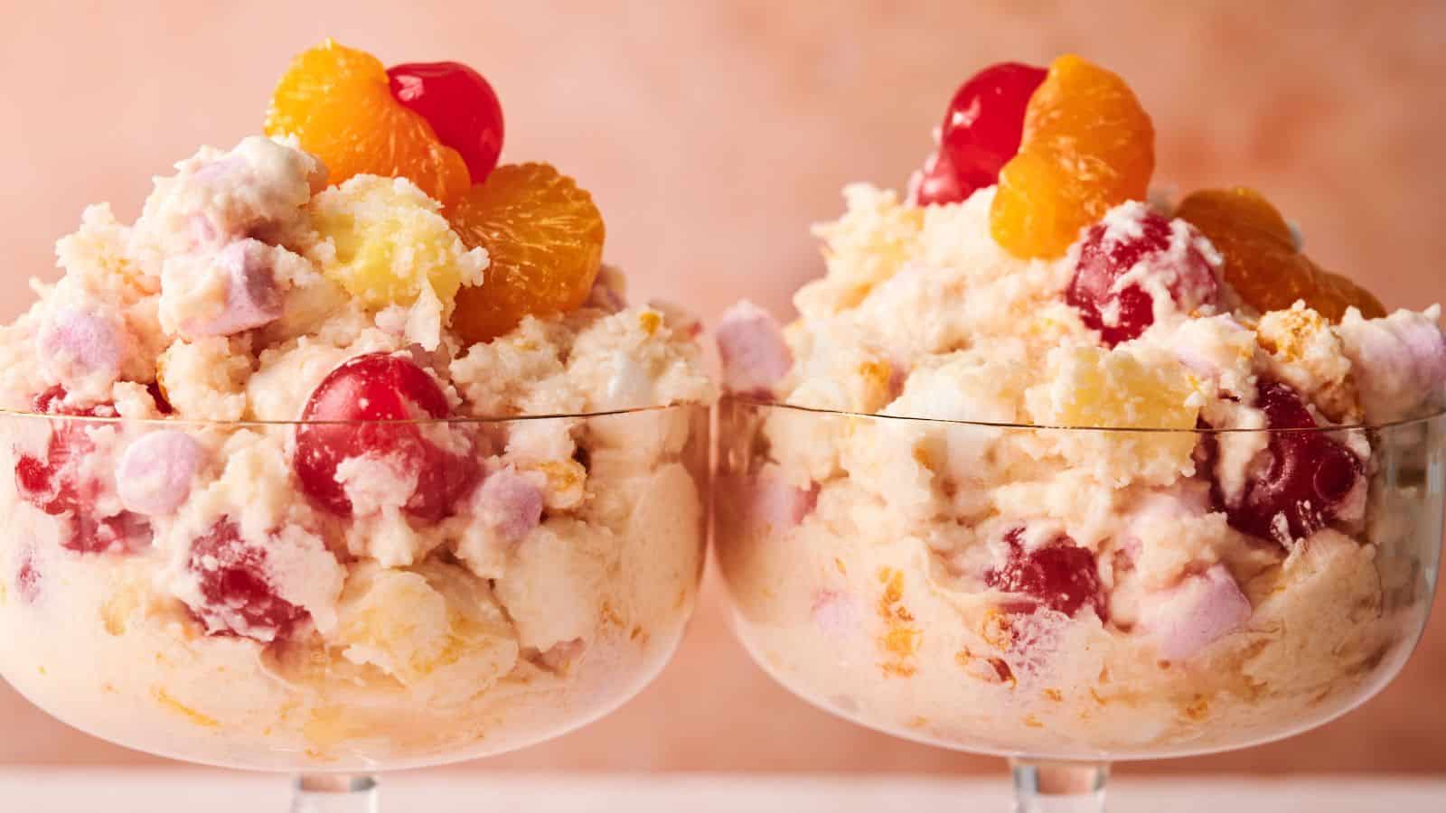 Two glass dessert cups filled with ambrosia salad, featuring whipped cream, cherries, pineapple, mandarin oranges, and marshmallows against a soft pink background.