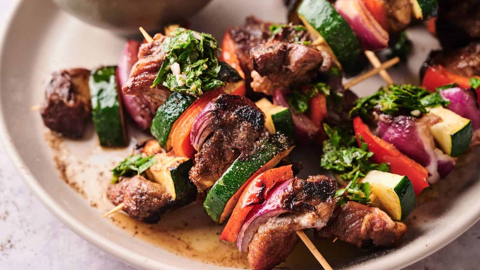 Grilled meat and vegetable skewers with zucchini, red onion, and red bell pepper, garnished with chopped herbs, served on a beige plate.