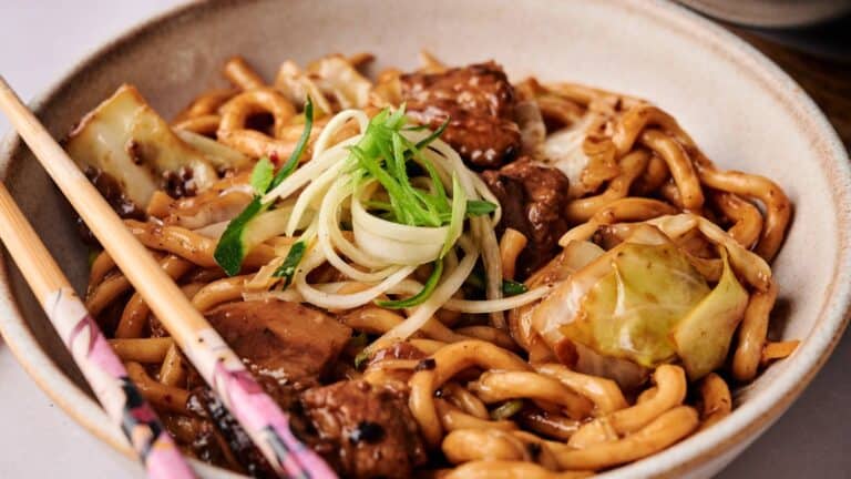 A bowl of stir-fried noodles with beef, cabbage, and sliced green onions, served with chopsticks.