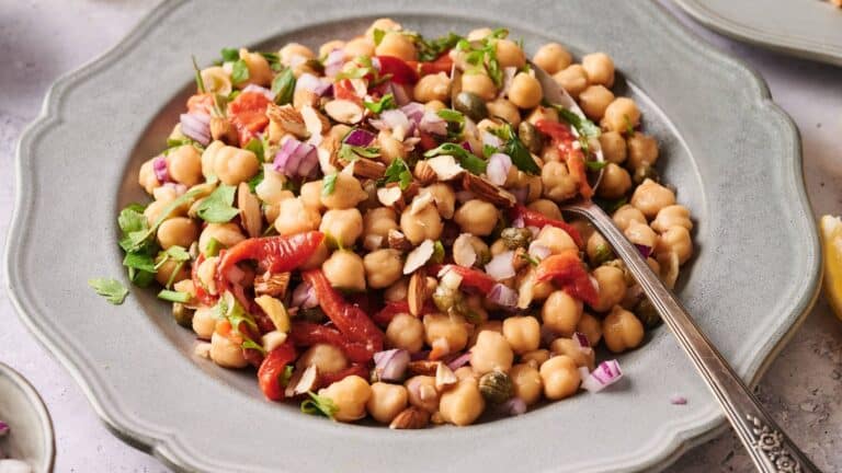 A plate of chickpea salad with chopped red onion, roasted red peppers, capers, herbs, and sliced almonds, served with a spoon.