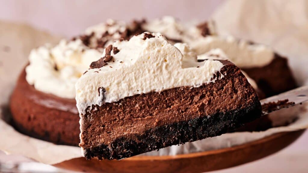 A slice of chocolate cheesecake with a dark crust and a thick layer of whipped cream is being lifted from the whole cake.