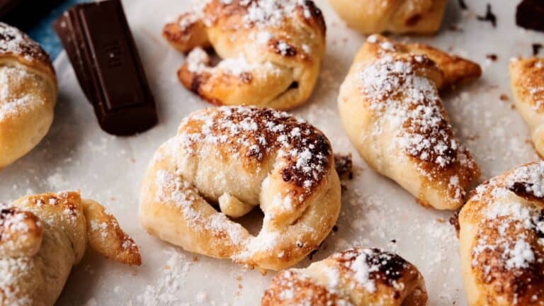 Close-up of several crescent-shaped pastries dusted with powdered sugar on parchment paper, with pieces of chocolate nearby.