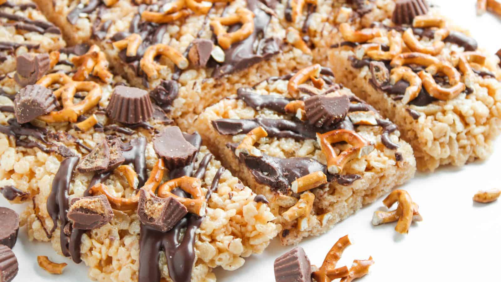 Rice crispy treat bars topped with drizzled chocolate, mini peanut butter cups, and crushed pretzels, cut into squares and arranged on a white surface.