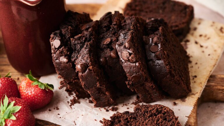 Sliced chocolate loaf cake with chocolate chips on parchment paper, next to fresh strawberries and a jar of chocolate sauce.
