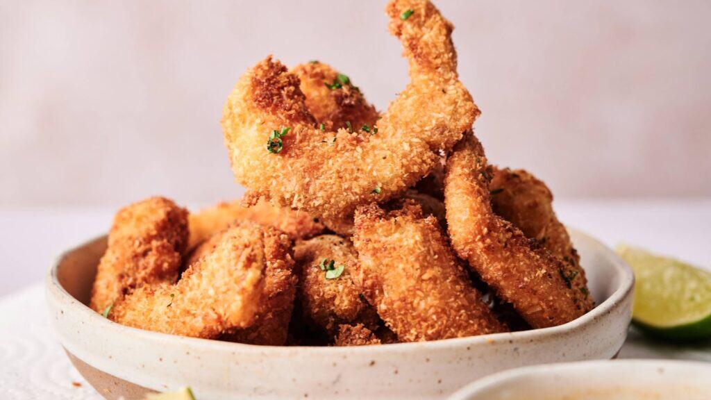 A bowl filled with golden-brown, breaded and fried shrimp garnished with chopped herbs, with a wedge of lime visible on the side.