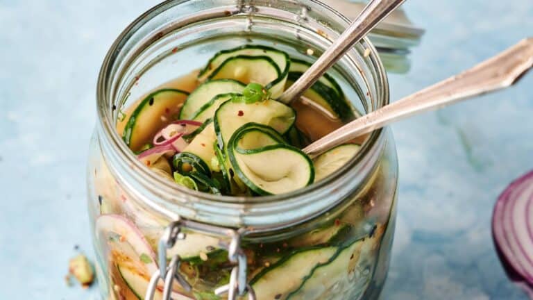 A glass jar filled with sliced cucumbers and red onions in brine, with two metal forks inside, set on a light blue surface.