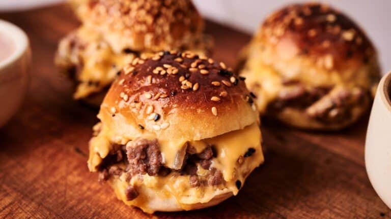 Three sesame seed slider buns filled with melted cheese and ground beef are arranged on a wooden surface.