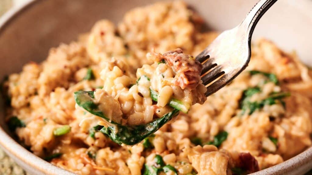 A close-up of a fork lifting a bite of creamy risotto with spinach and pieces of tomato from a bowl.