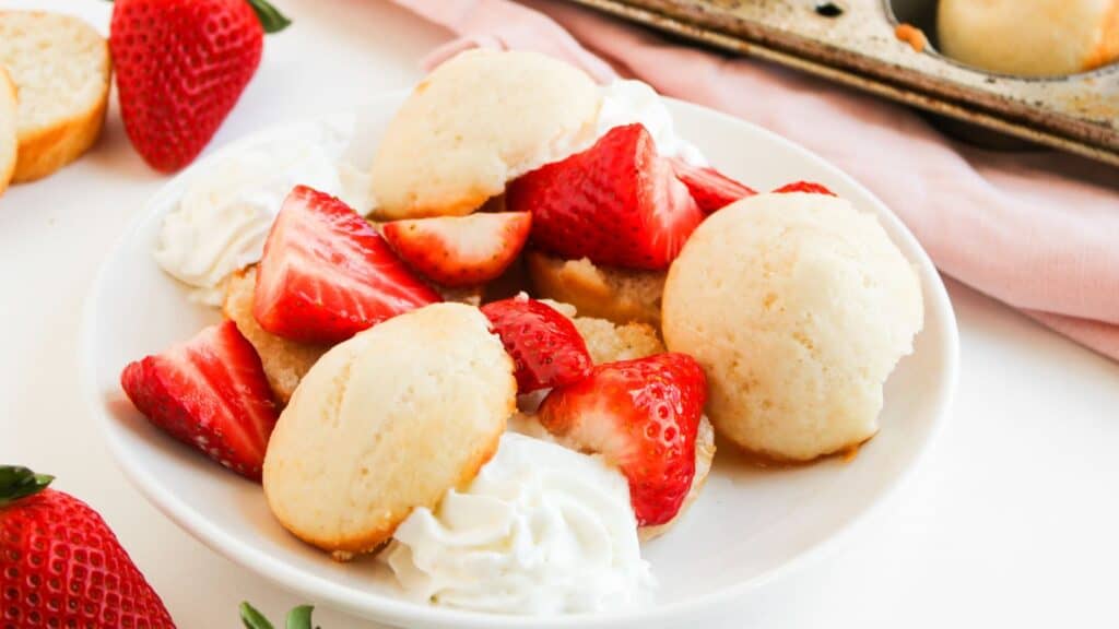 A plate with sliced strawberries, dollops of whipped cream, and several shortcake biscuits. A muffin tin and whole strawberries are visible in the background.