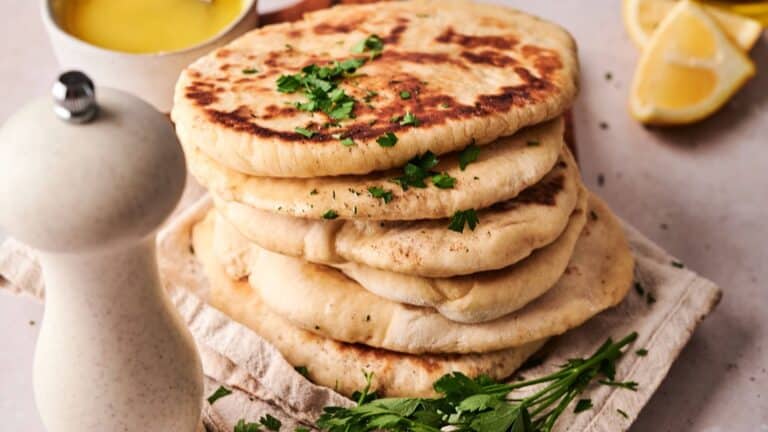 A stack of flatbreads garnished with chopped parsley sits on a cloth, with a pepper grinder, fresh herbs, a cup of yellow sauce, and lemon wedges nearby.