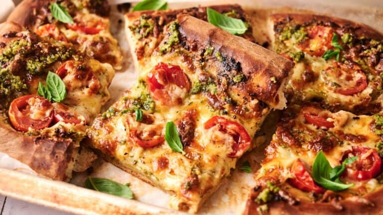 Rectangular pizza slices topped with melted cheese, cherry tomatoes, pesto, and fresh basil leaves on a baking tray.