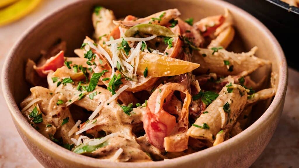 A bowl of penne pasta with creamy sauce, sliced bell peppers, grated cheese, and chopped parsley.