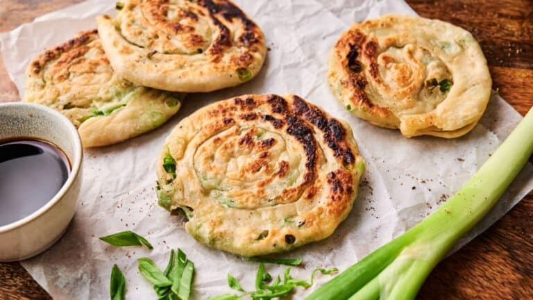 Four cooked scallion pancakes on parchment paper with sliced green onions and a small dish of dipping sauce.