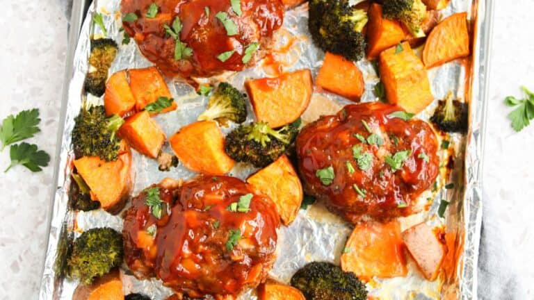 Sheet pan with three glazed meatloaf portions, roasted sweet potato chunks, and broccoli florets, garnished with chopped herbs.