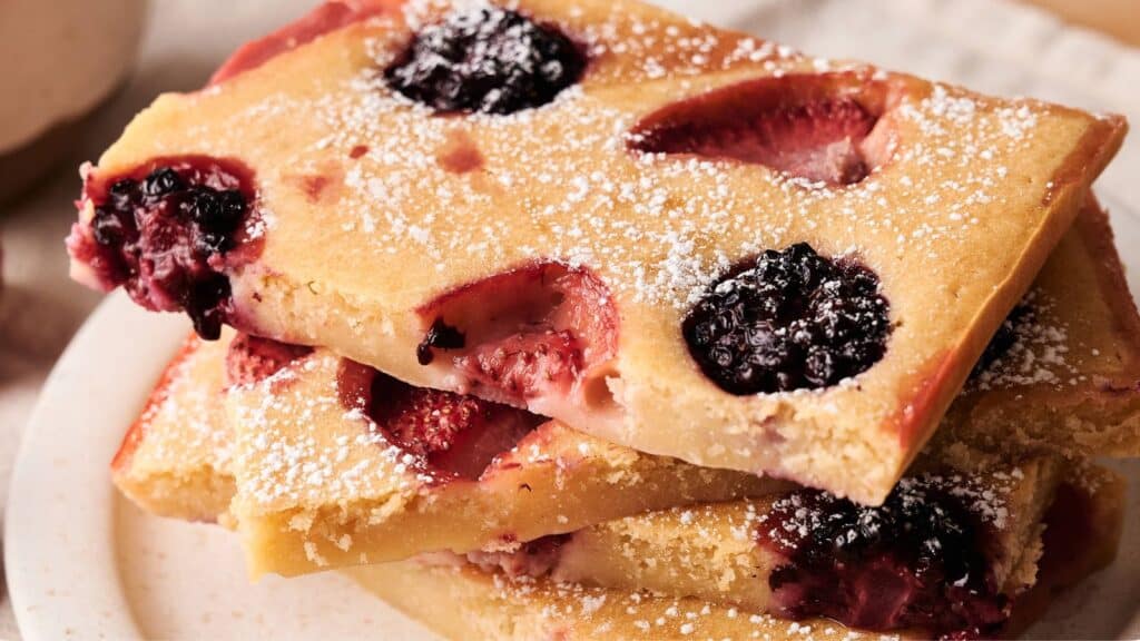 Three rectangular slices of baked fruit-studded cake, topped with powdered sugar, are stacked on a white plate.
