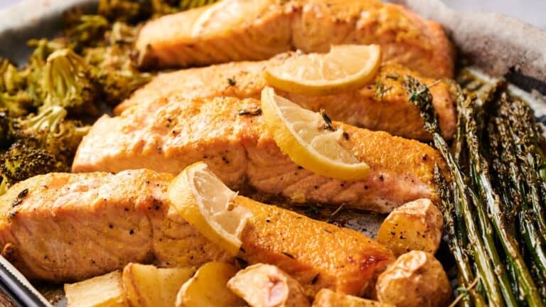 Four baked salmon fillets garnished with lemon slices, surrounded by roasted broccoli, asparagus, potatoes, and garlic cloves on a sheet pan.