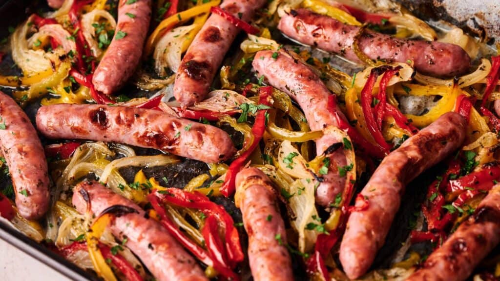 Sheet pan with roasted sausages, sliced red and yellow bell peppers, and onions, garnished with chopped parsley.