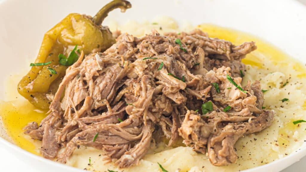 Shredded beef served over mashed potatoes, garnished with a pepper and fresh herbs, with broth pooled around the dish.