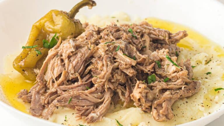 Shredded beef served over mashed potatoes, garnished with a pepper and fresh herbs, with broth pooled around the dish.